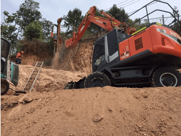 An Orange Hitachi Excavator undertaking construction drilling into dirt with Beards team member standing by